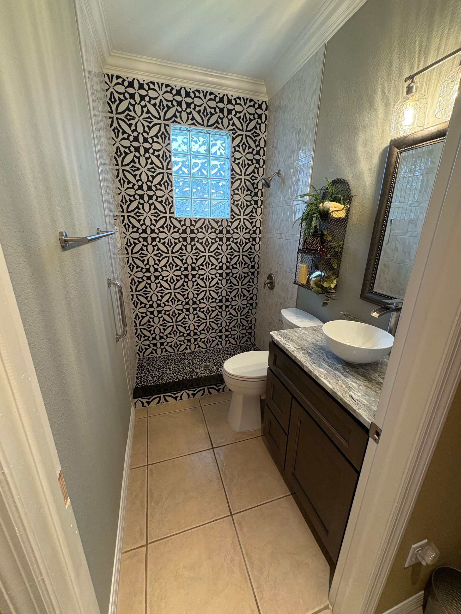 Narrow bathroom with patterned shower curtain, toilet, dark vanity, round mirror, and beige tile floor.