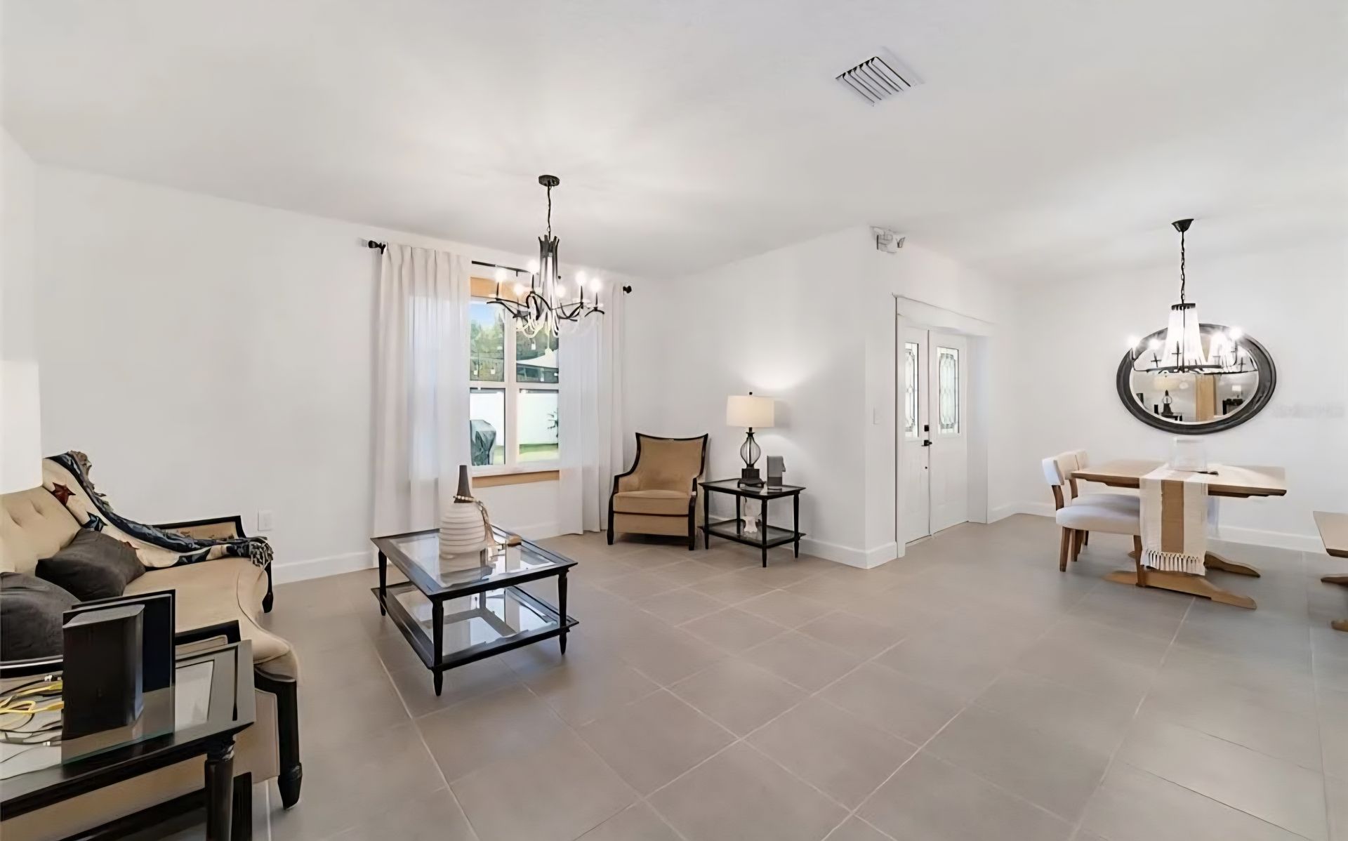 Bright open living and dining room with gray tile floor, white walls, chandelier, and large windows.