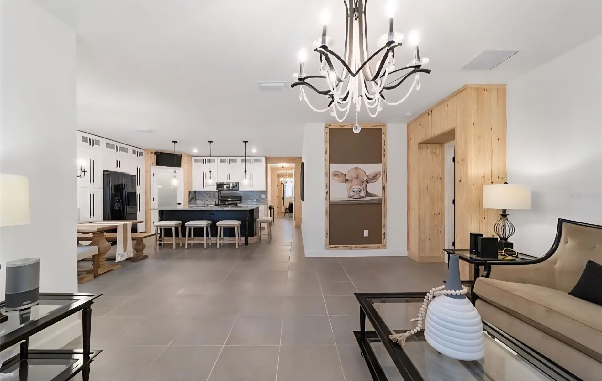 Spacious modern living room with gray tile floors, chandelier, beige sofa, and open kitchen in the background