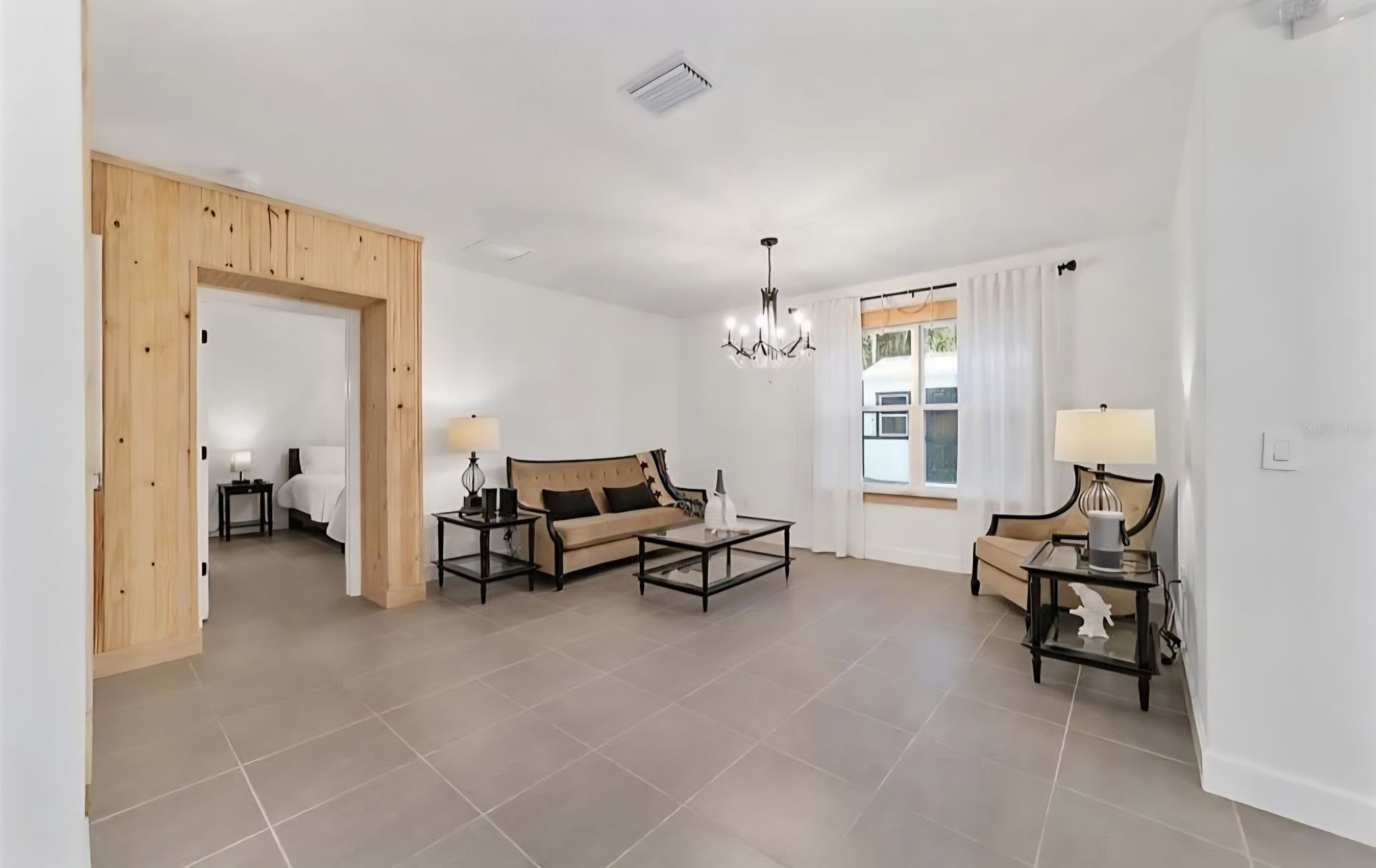 Bright living room with tiled floor, beige sofa, chandelier, chair, lamp, and large window.