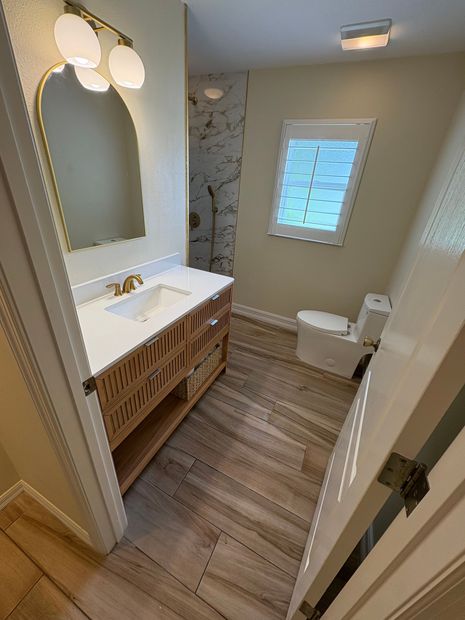 Small bathroom with white vanity, oval mirror, wood cabinet, window, and toilet.