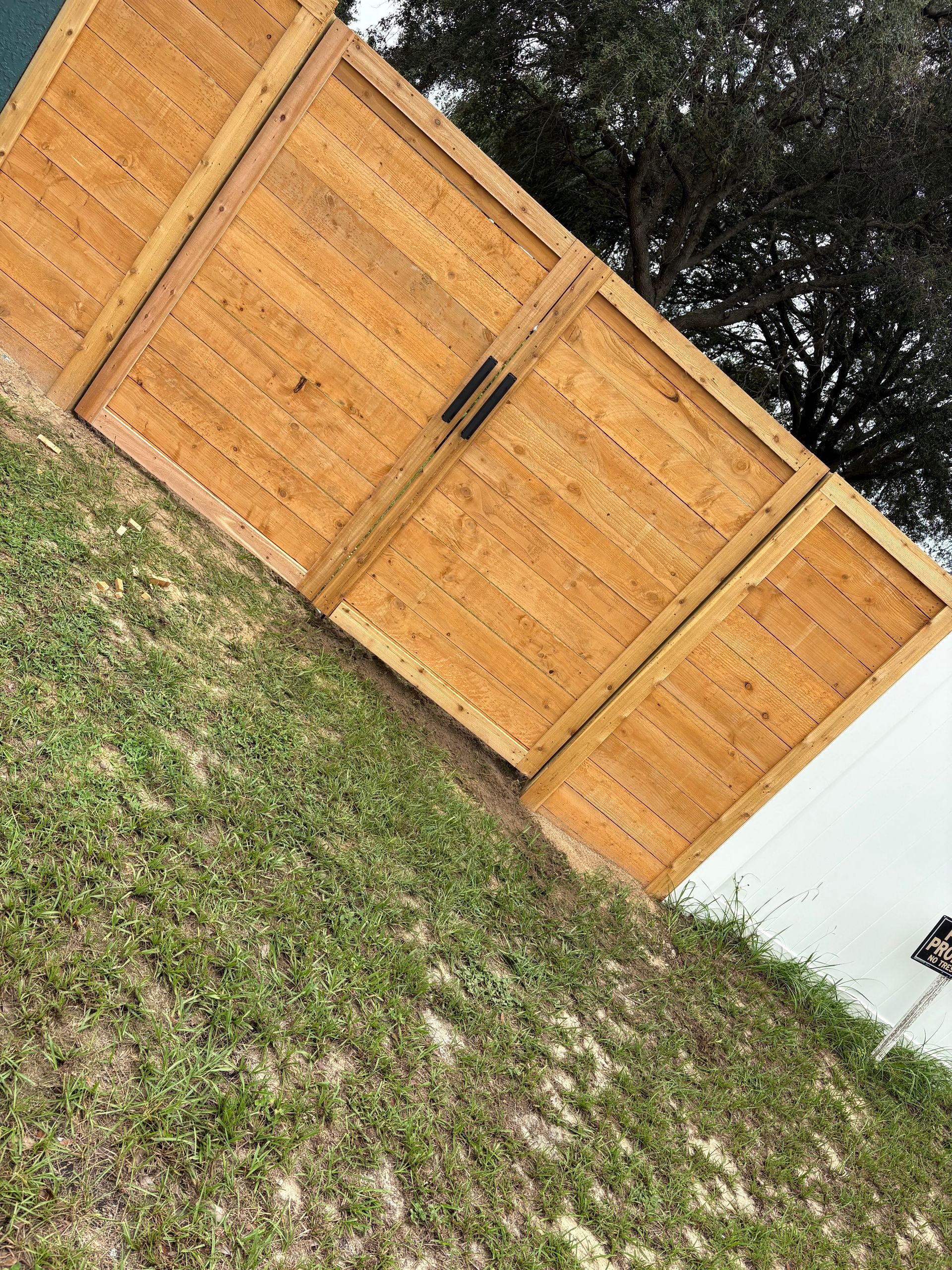 Wooden backyard fence with gate, beside a white wall and patchy grass lawn.