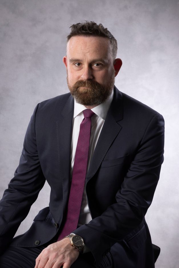 Portrait of a bearded man in a dark suit and magenta tie, seated against a gray studio background