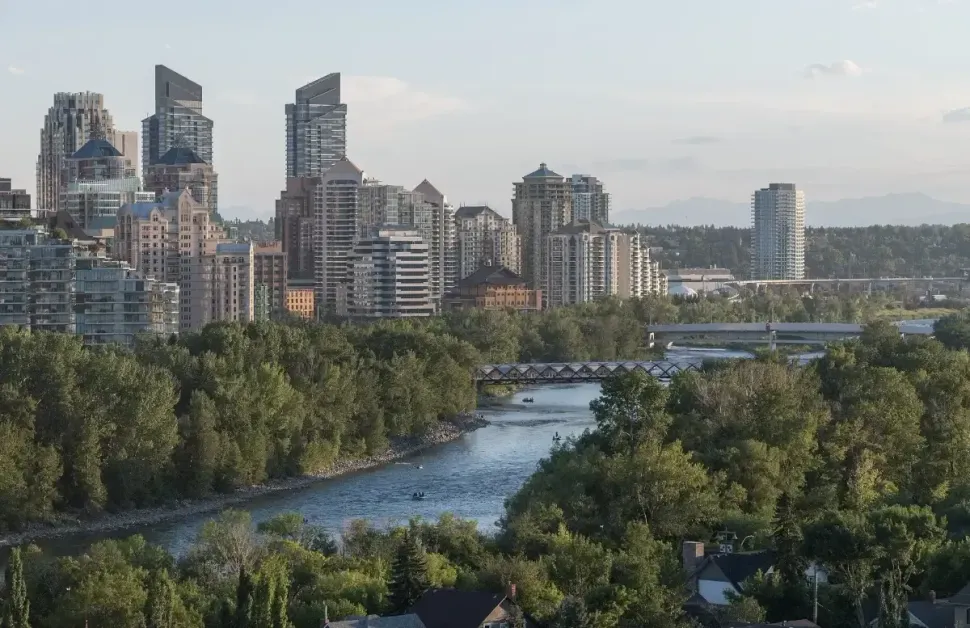City skyline beside a river with trees and distant hills under a clear sky