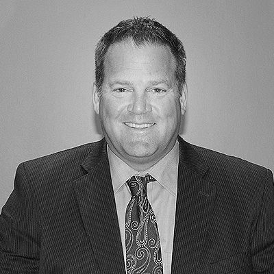 A professional headshot of a smiling person wearing a suit, a collared shirt, and a patterned tie against a plain wall.