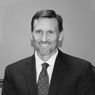 A man smiling in a dark suit and patterned tie, set against a plain background.