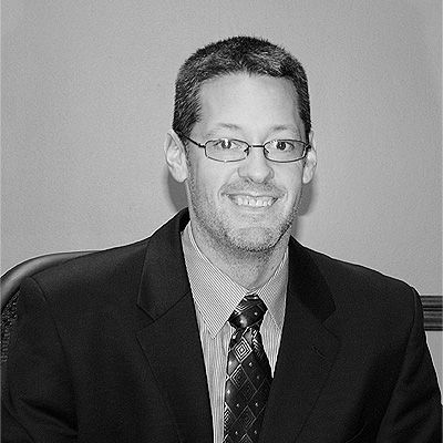 A person with short hair and glasses wearing a suit and tie, smiling against a plain background.