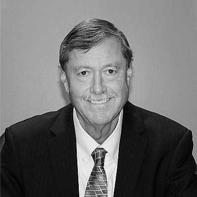 A professional headshot of a man with a mustache, wearing a suit and patterned tie, smiling against a plain background.