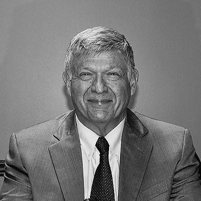 A professional headshot of a person in a suit and tie, smiling slightly against a plain gray background.