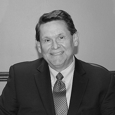 A headshot of a smiling person wearing a dark suit, light-colored shirt, and a patterned tie, set against a plain background.