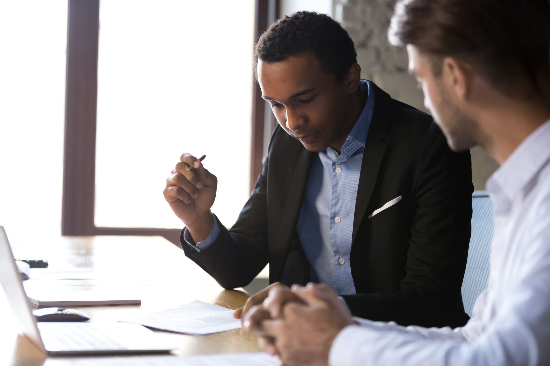 Two professionals sit at a desk, reviewing documents and discussing work in a bright, modern office setting.