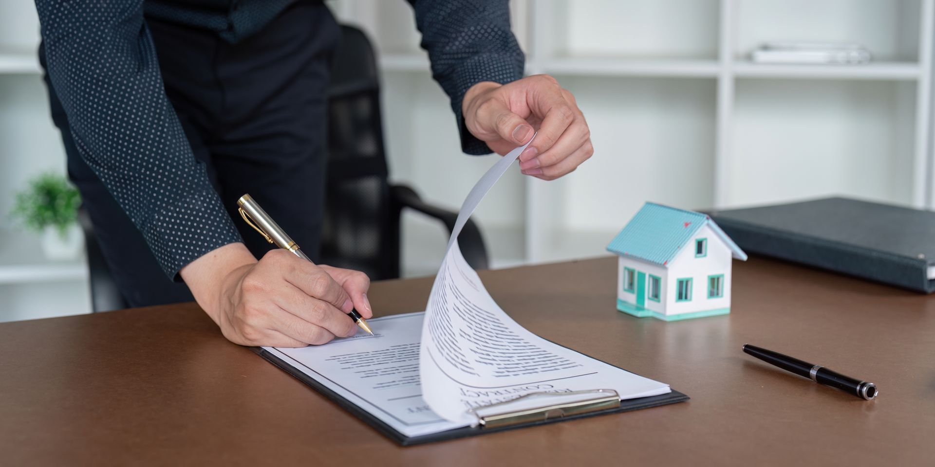 A person in a dark shirt signs a document at a desk next to a miniature house model and a pen.