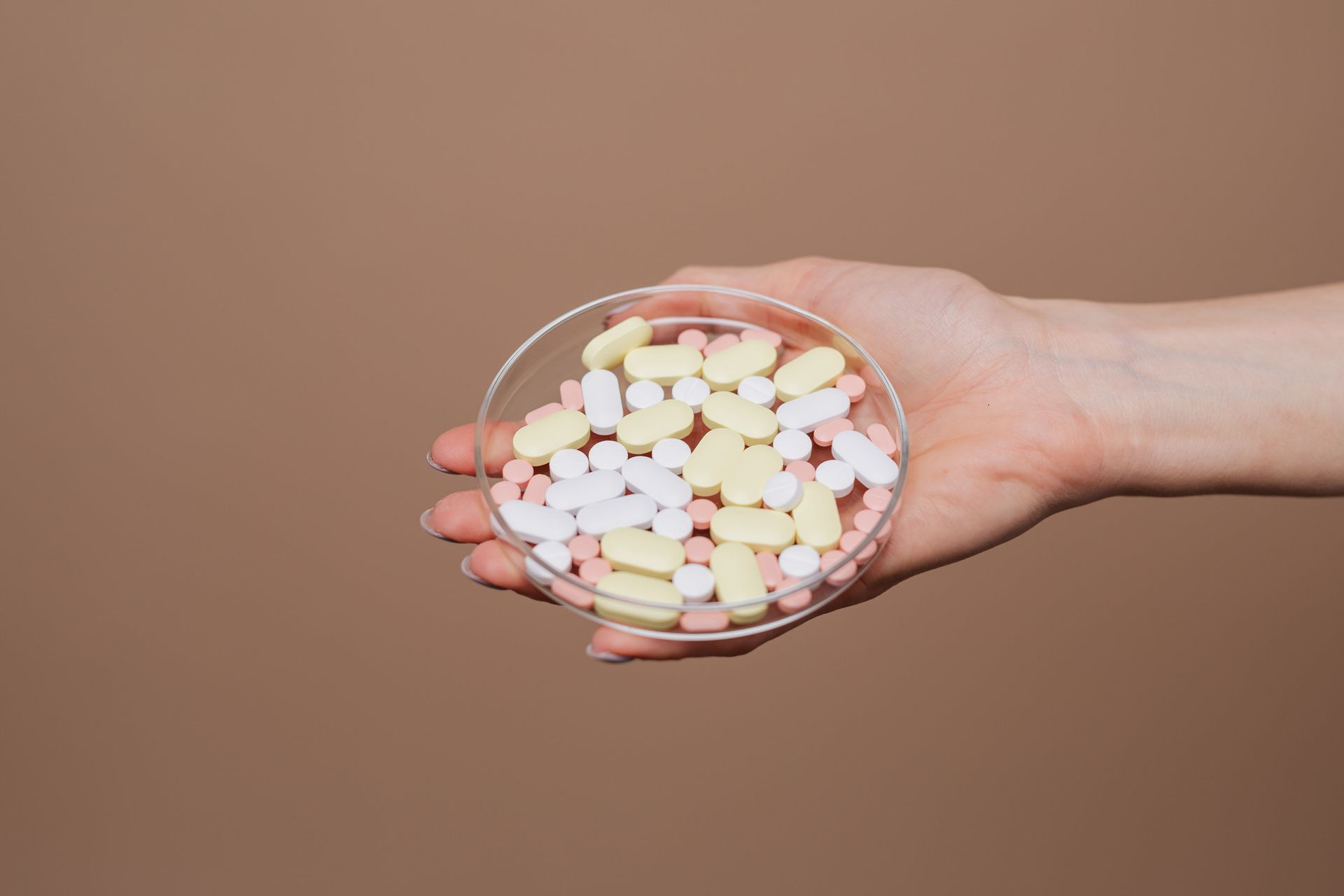 Hand holding a clear dish of assorted pastel pills on a beige background
