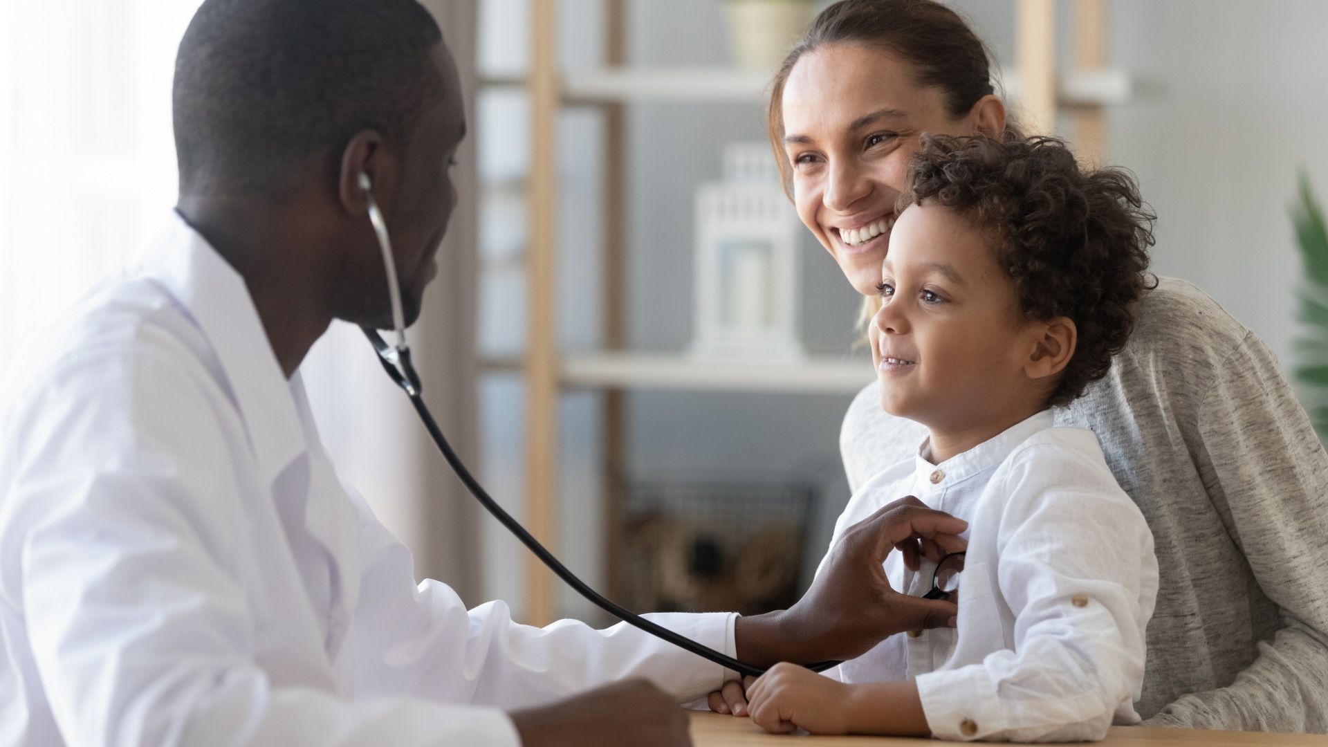 Doctor checking a child's heartbeat with a stethoscope while parent smiles nearby in a clinic