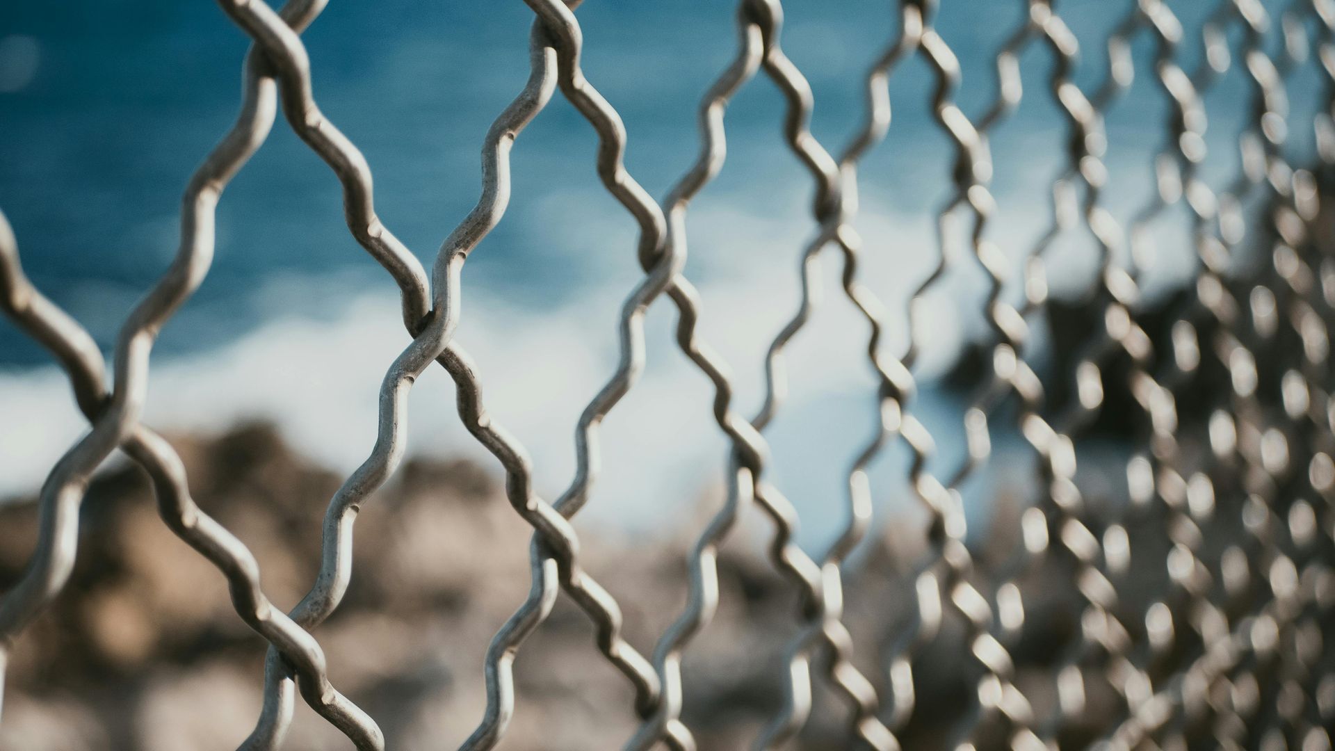 Close-up of a metal chain-link fence with a blurred view of the ocean and sky.