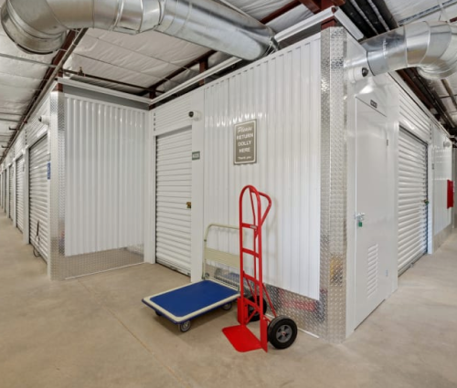 White storage units with roll-up doors in a warehouse, with a red hand truck and blue dolly in front.