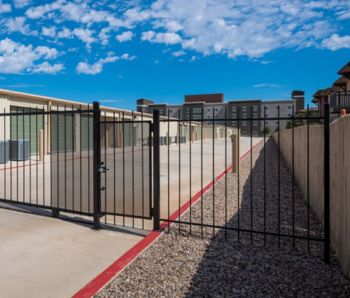 Gated outdoor walkway with black metal fence, concrete path, red curb, and storage facility under a blue sky