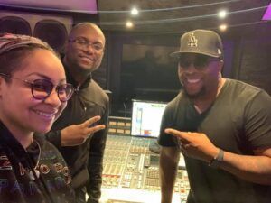 Three people smiling in a recording studio, posing beside an audio mixing console.