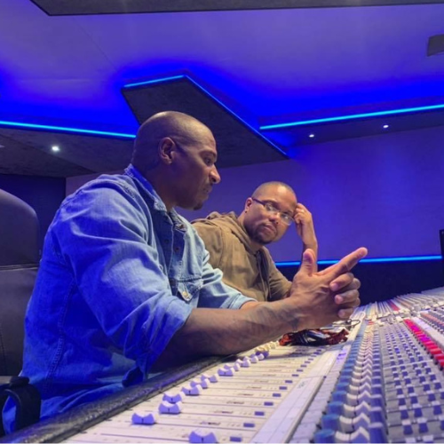 Two people in a blue-lit recording studio, seated at a mixing console and discussing audio settings.