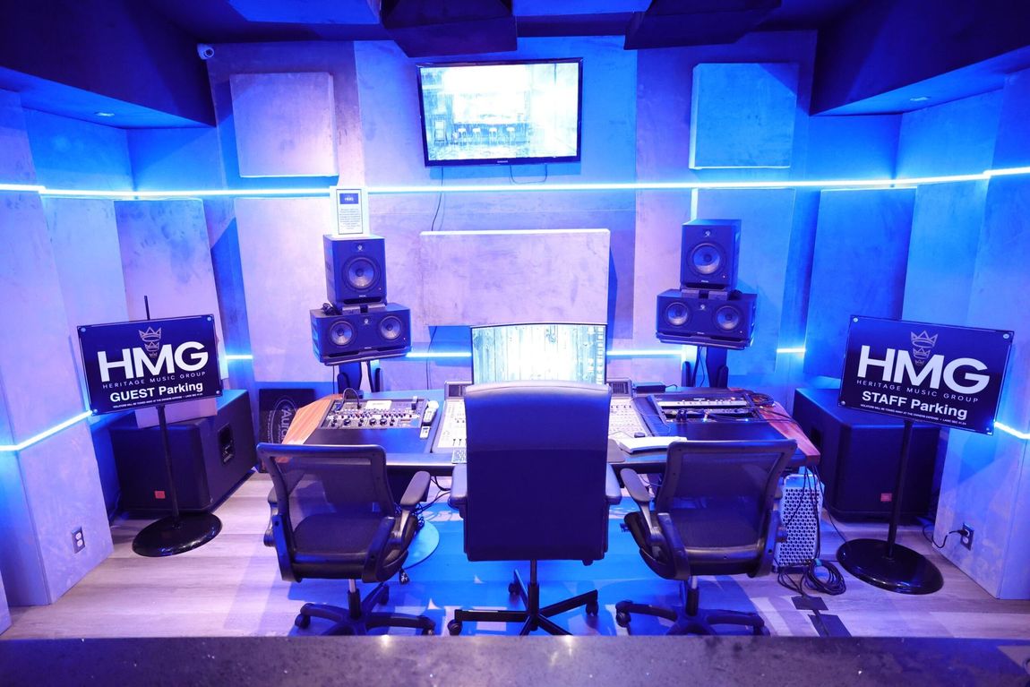 Blue-lit music studio with mixing desk, monitors, speakers, and two HMG signs on either side.