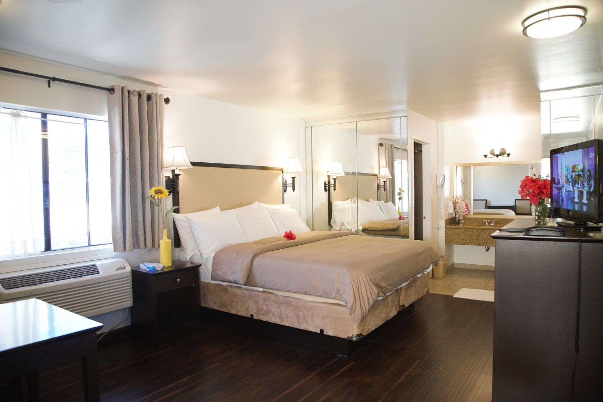 Bedroom with a queen bed, TV, and a bathroom visible in the mirror. Dark wood floor and white walls.