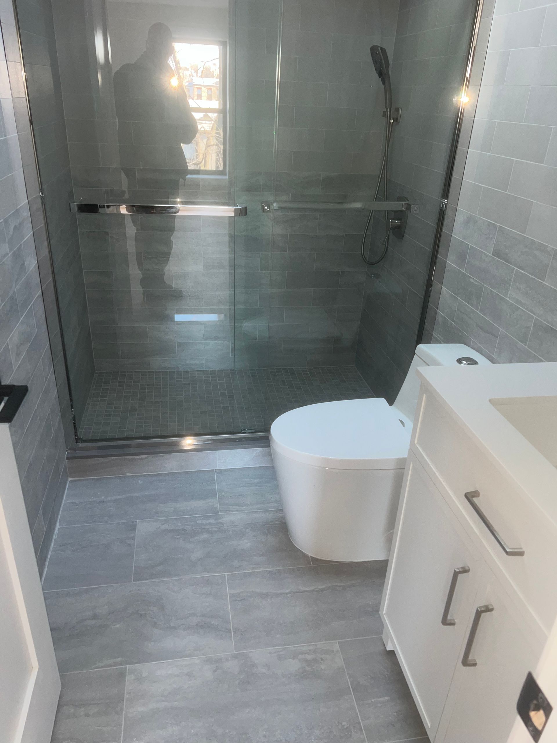 Modern bathroom with glass shower, white cabinet, and toilet on gray tile floor