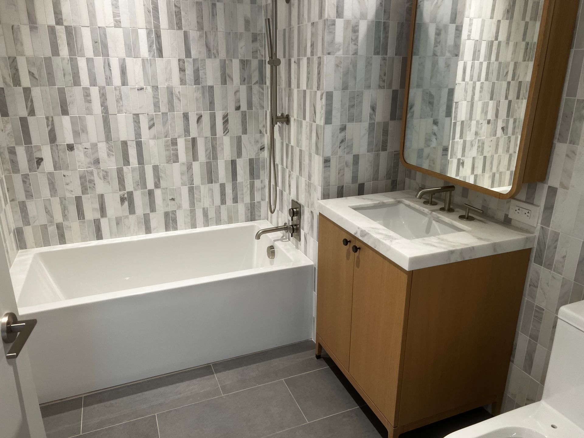 Small bathroom with a white tub, tiled walls, gray floor, and wooden sink vanity with mirror.