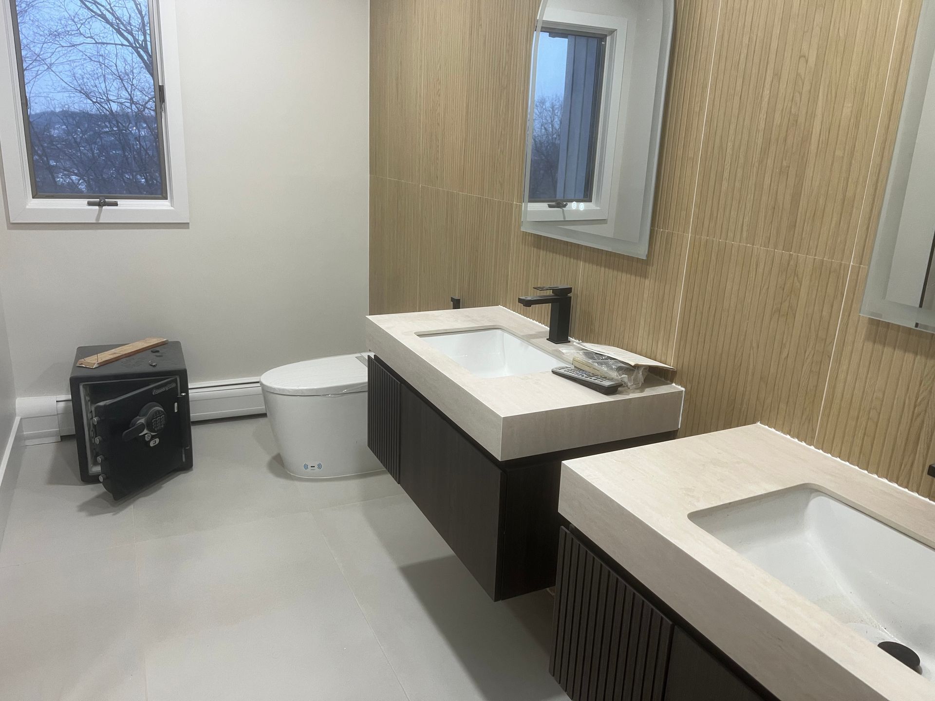 Modern bathroom with double sinks, mirrors, wood accent wall, and toilet near a window.