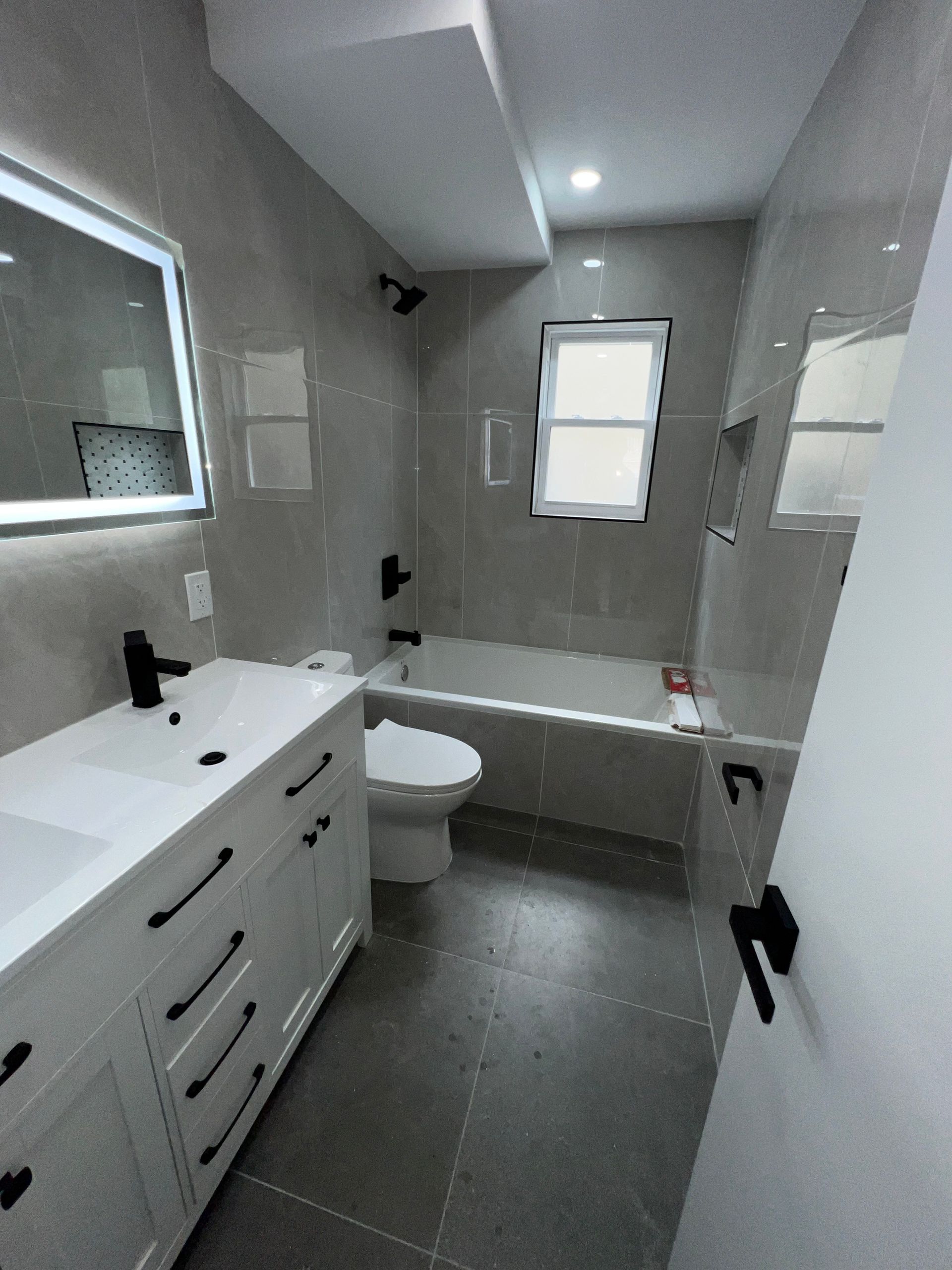 Modern white bathroom with vanity, toilet, bathtub, gray floor tiles, and a small window.
