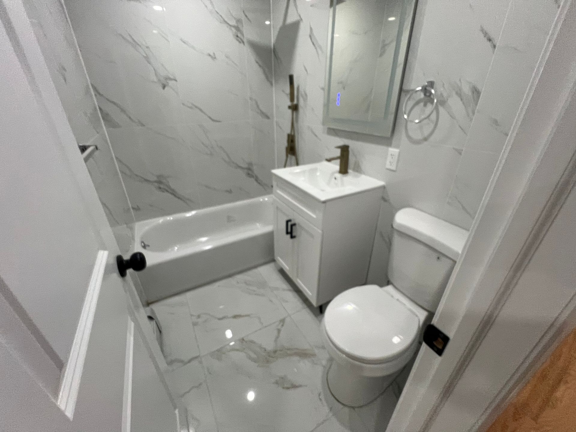 Small white bathroom with marble tile, sink cabinet, toilet, tub, and wall mirror.