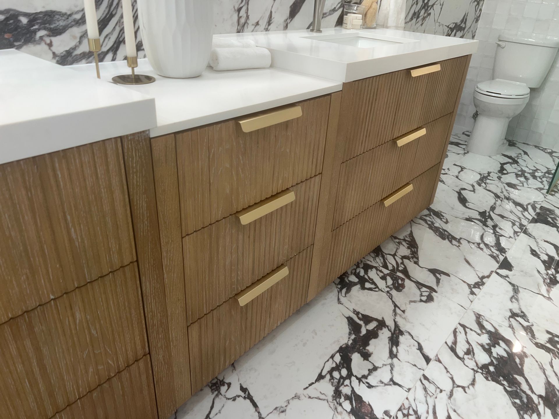 Wood vanity with white countertop and gold handles in a marble-tiled bathroom