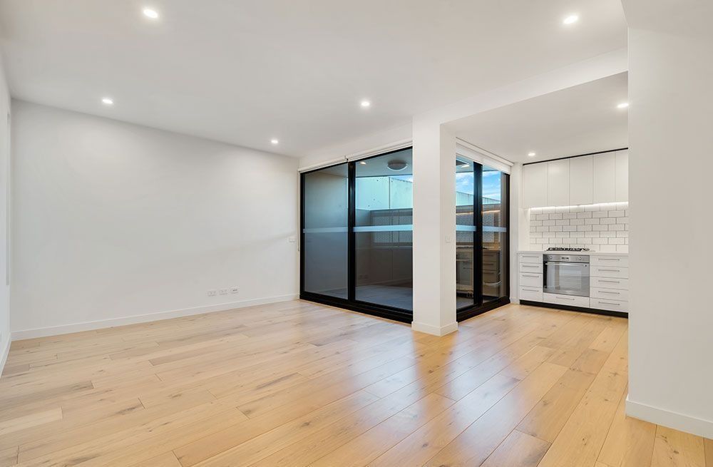 Bright empty modern apartment room with light wood floors, white walls, and kitchen area by sliding glass doors
