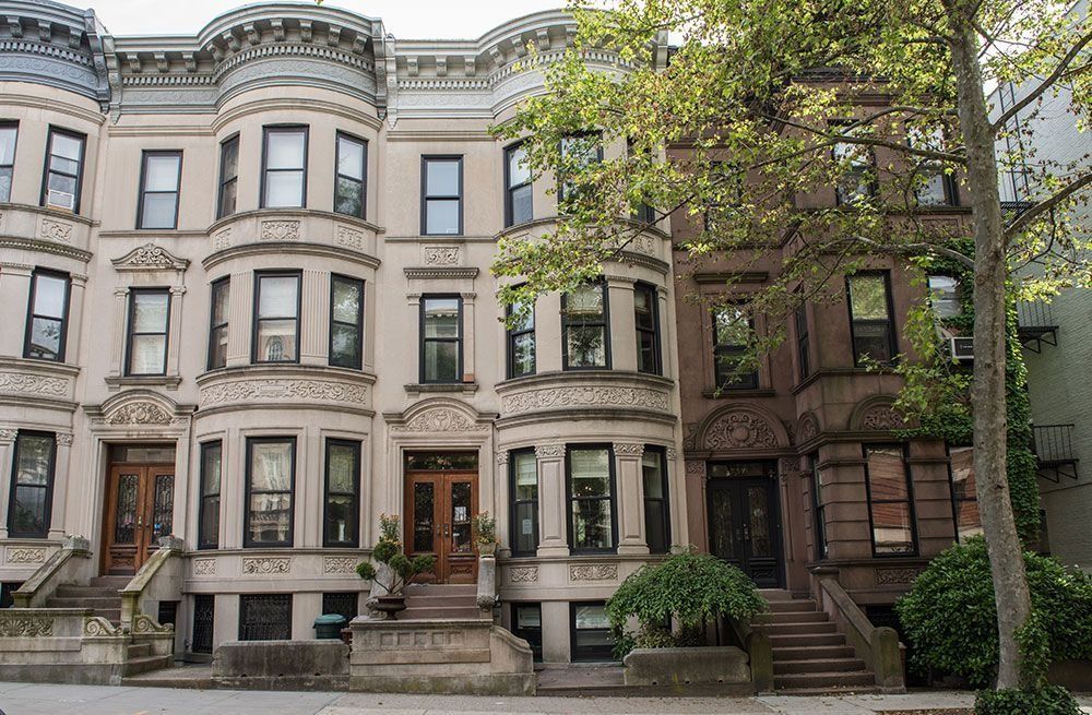 Row of brownstone apartment buildings with bay windows and trees along a quiet city street