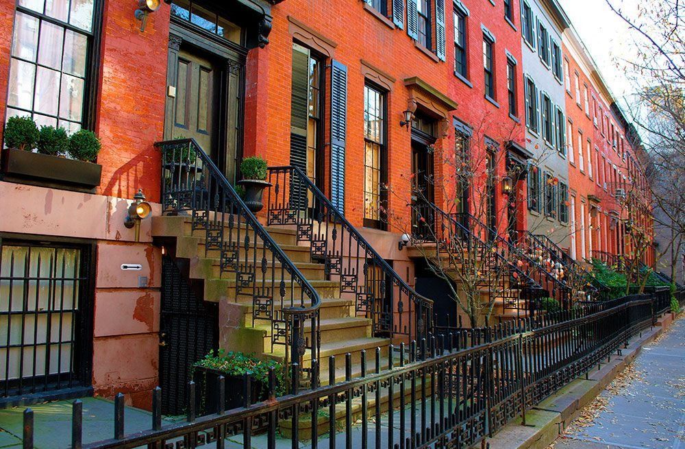 Row of red brick townhouses with black iron stoops and railings along a tree-lined street