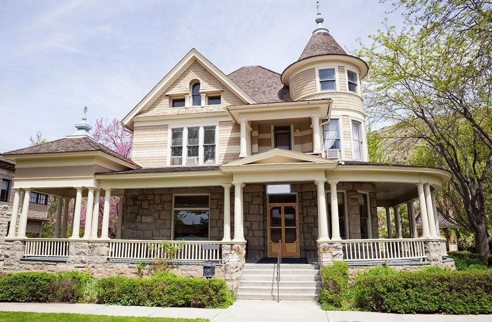 Victorian-style house with a wraparound porch, turret, and beige exterior surrounded by greenery