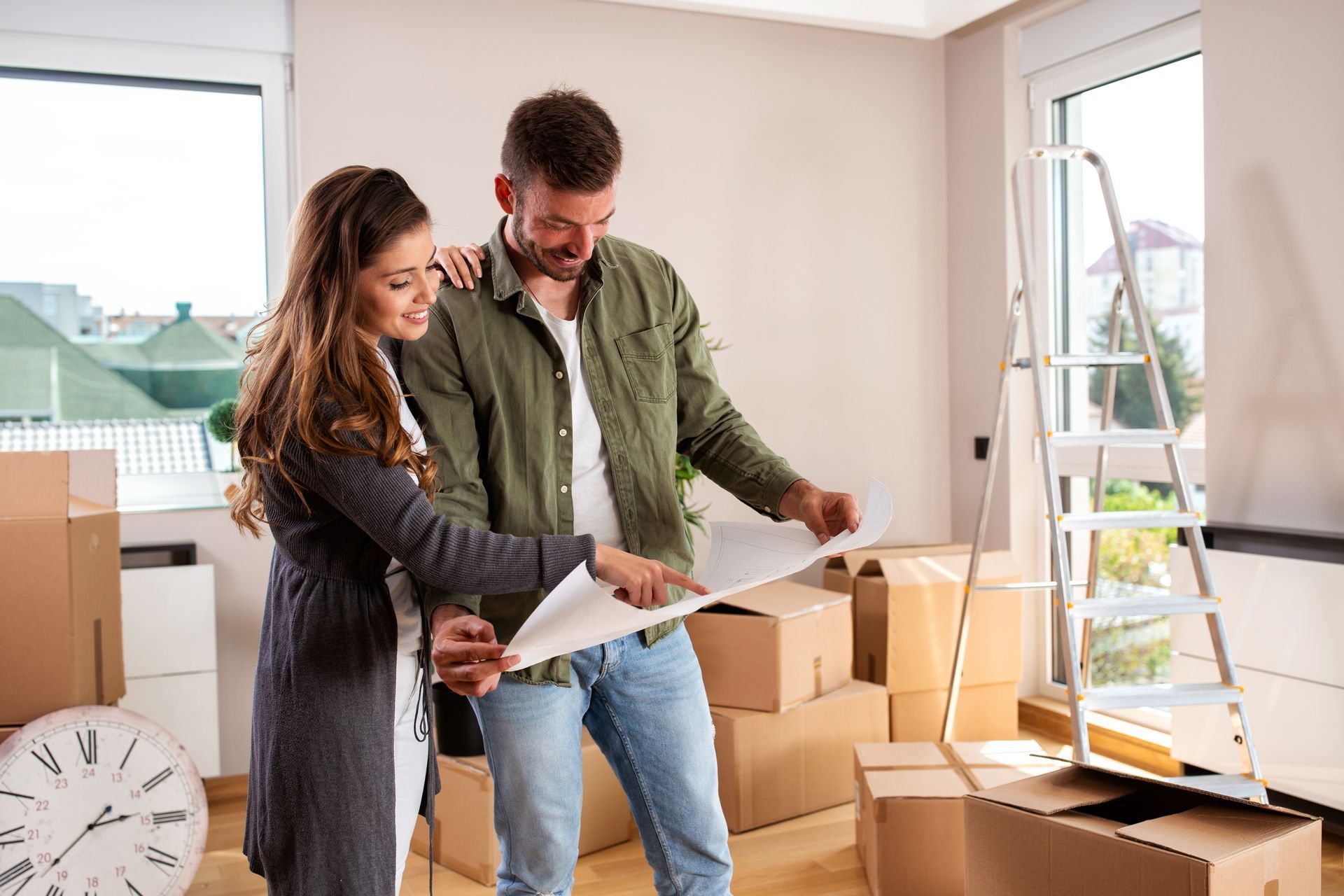 Couple reviewing a floor plan in a moving room surrounded by cardboard boxes and a ladder