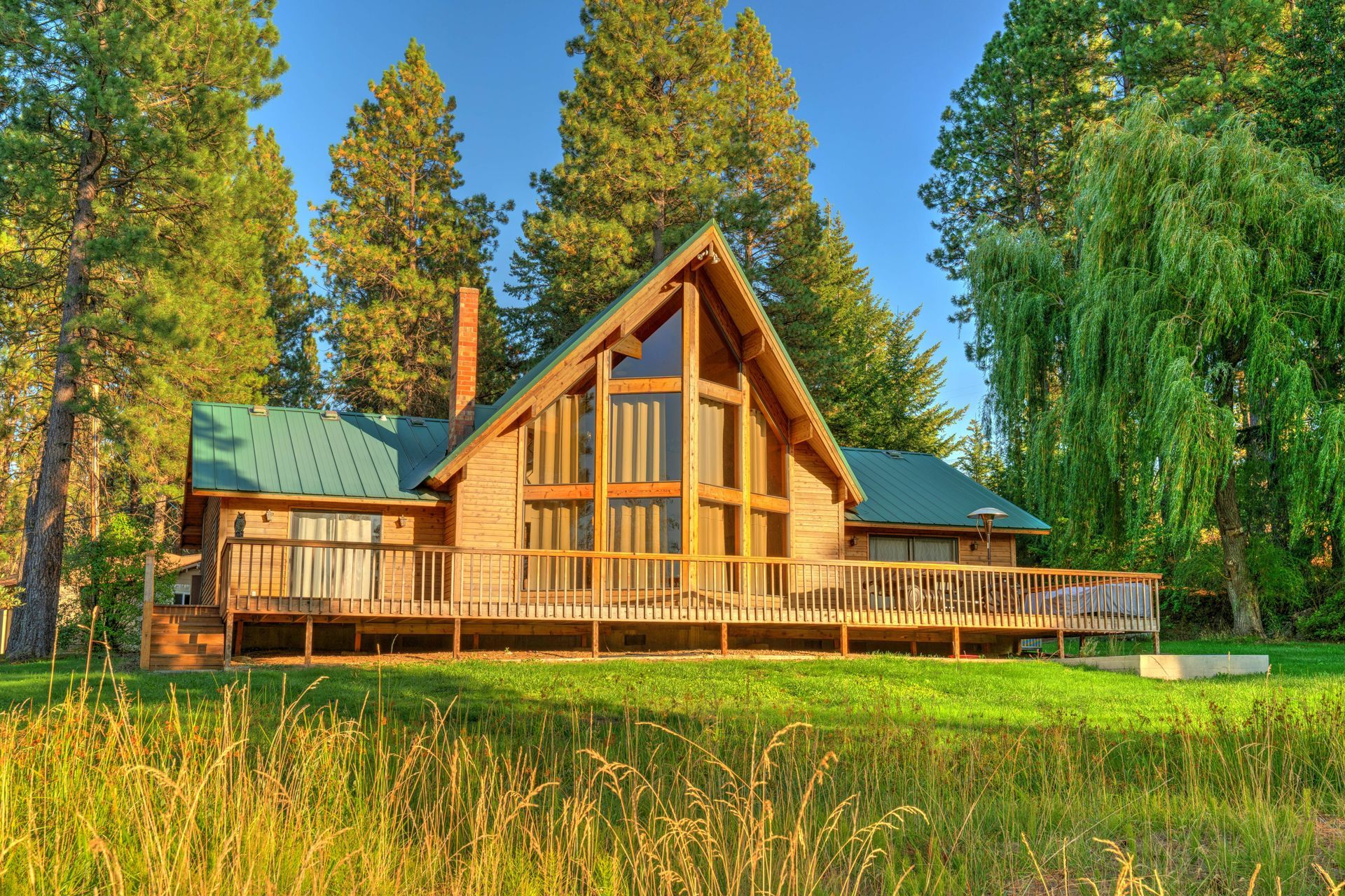 Wooden cabin with a steep roof in a sunlit forest clearing, surrounded by tall trees and grass.