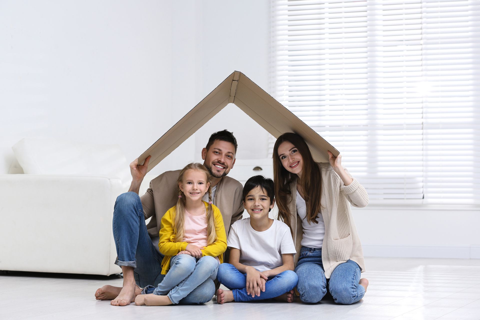 Family sitting inside a cardboard house frame in a bright living room, smiling at the camera