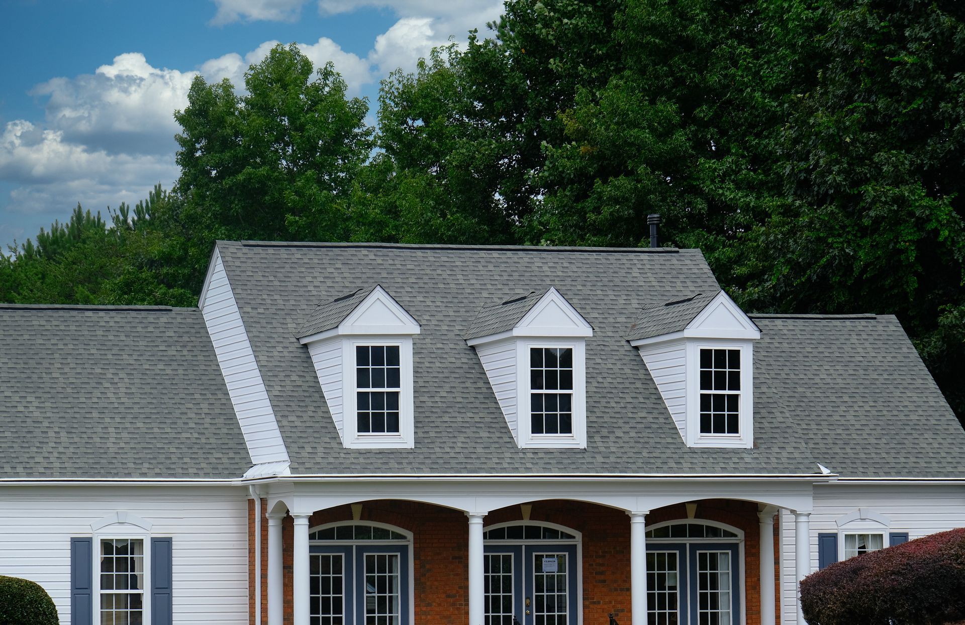 White house roof with dormer windows against green trees and a cloudy blue sky