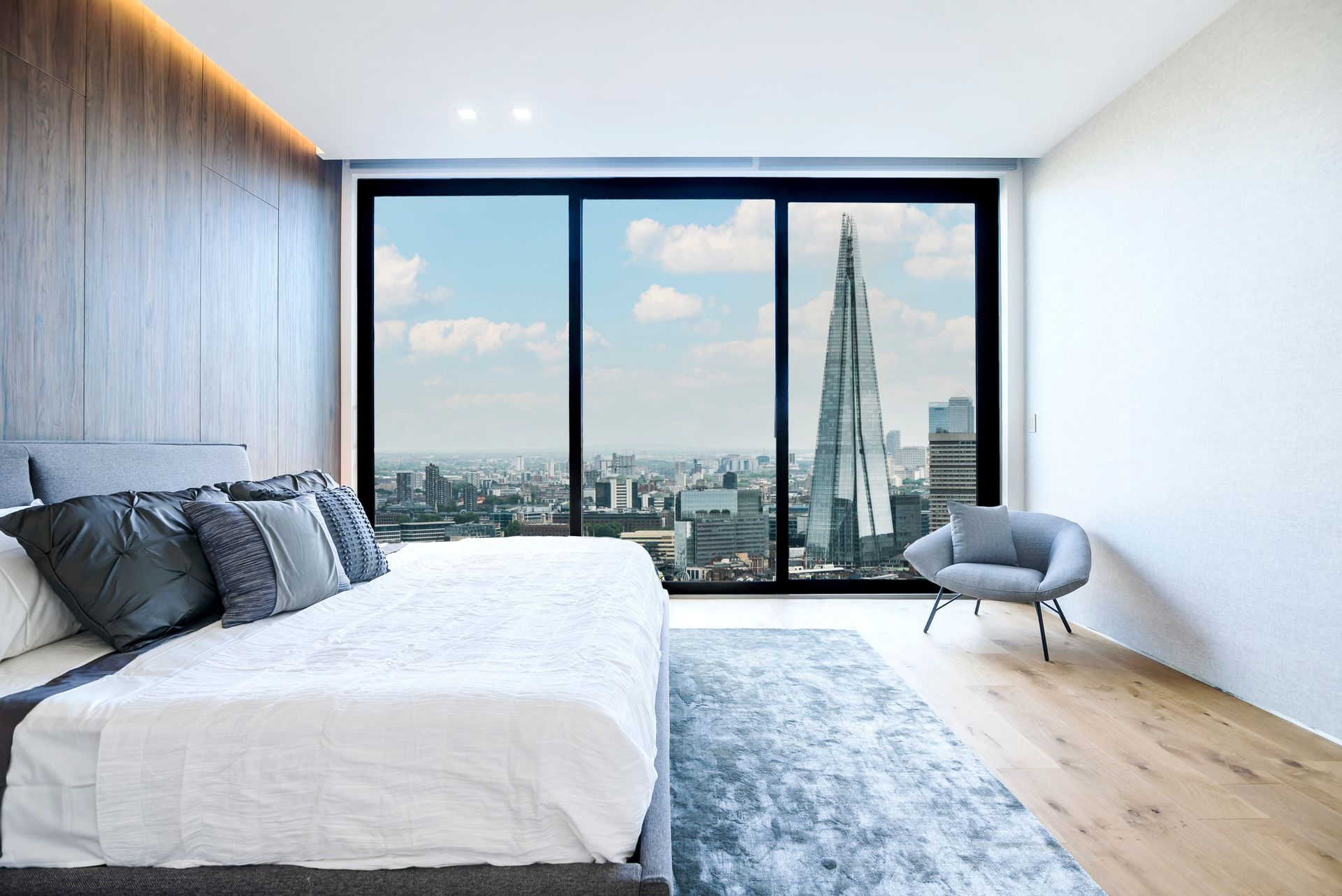 Modern bedroom with a large window, city skyline view, white bed, blue rug, and gray chair