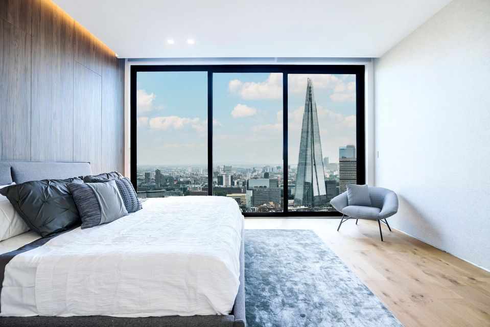 Modern bedroom with a large window, city skyline view, white bed, blue rug, and gray chair