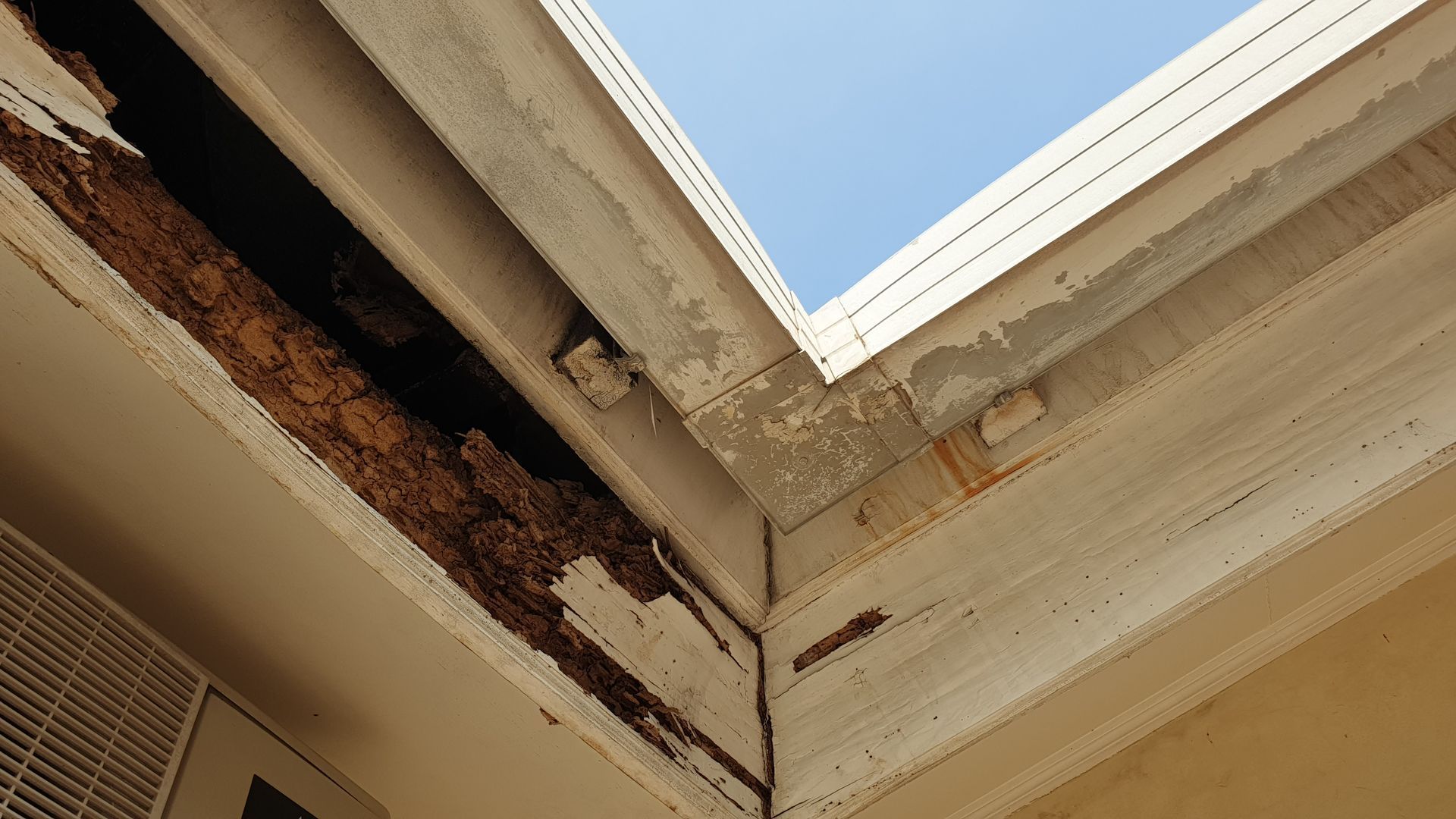 Damaged wooden roof corner with peeling paint and exposed rot against a blue sky