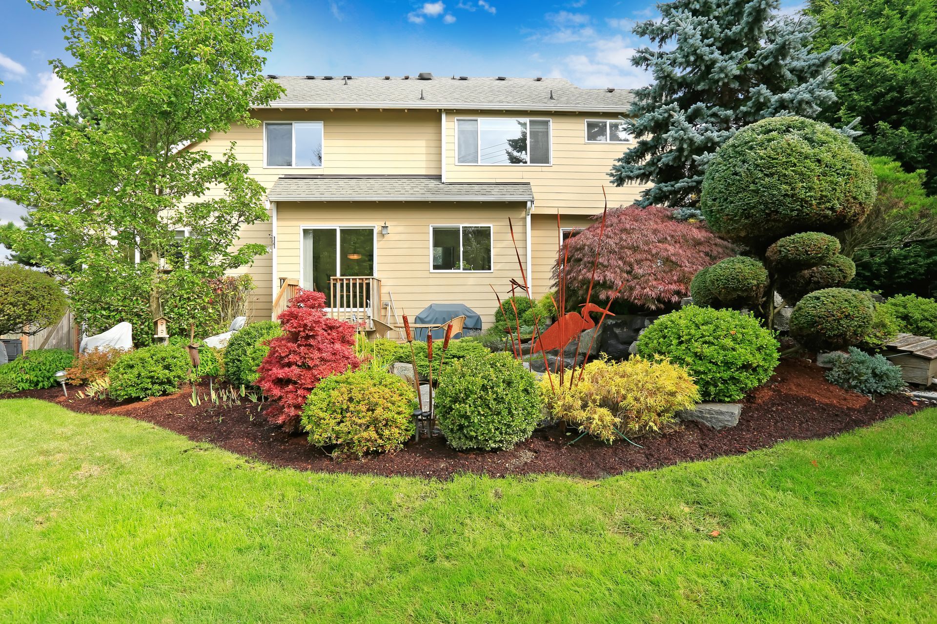 Two-story beige house with a manicured garden, colorful shrubs, and a green lawn