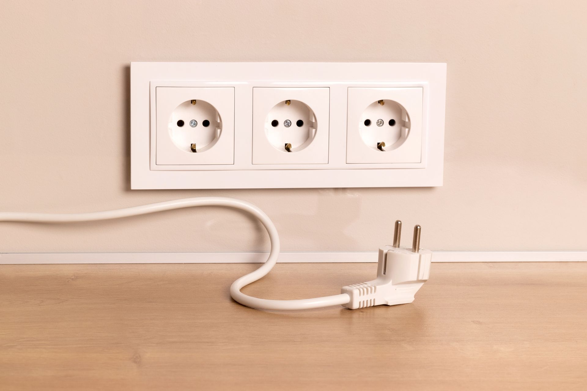 White wall outlet with a plugged-in cord on a wooden surface against a beige wall.