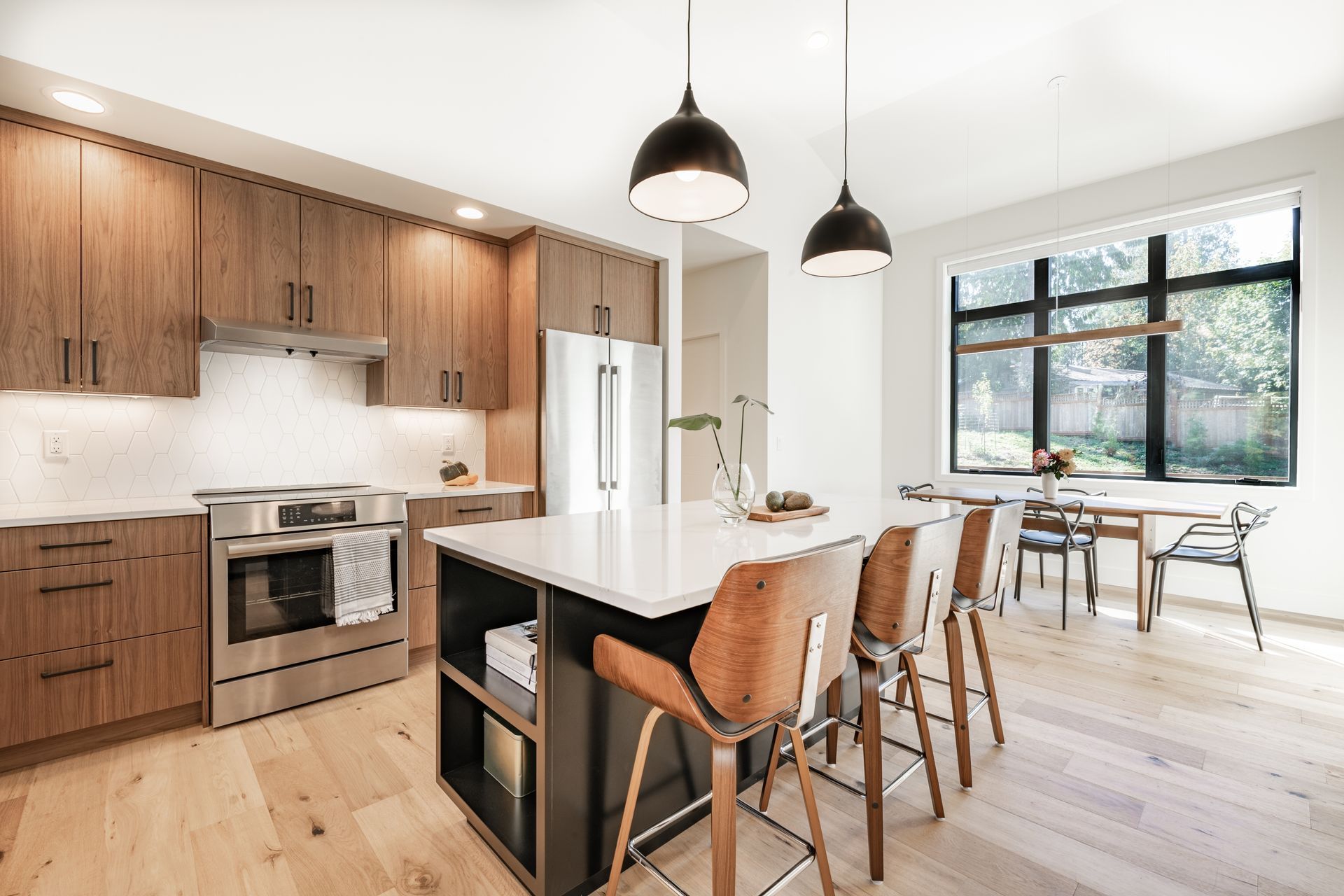 Modern open kitchen and dining area with wood cabinets, island stools, and large windows overlooking greenery