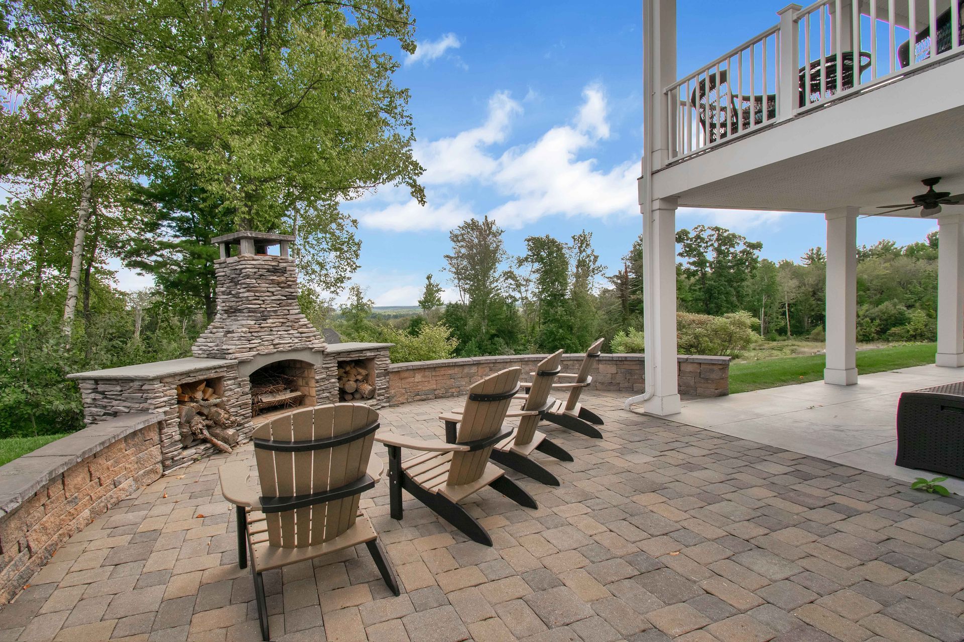 Stone patio with fireplace, chairs, and barrel table beside a house deck in a wooded yard
