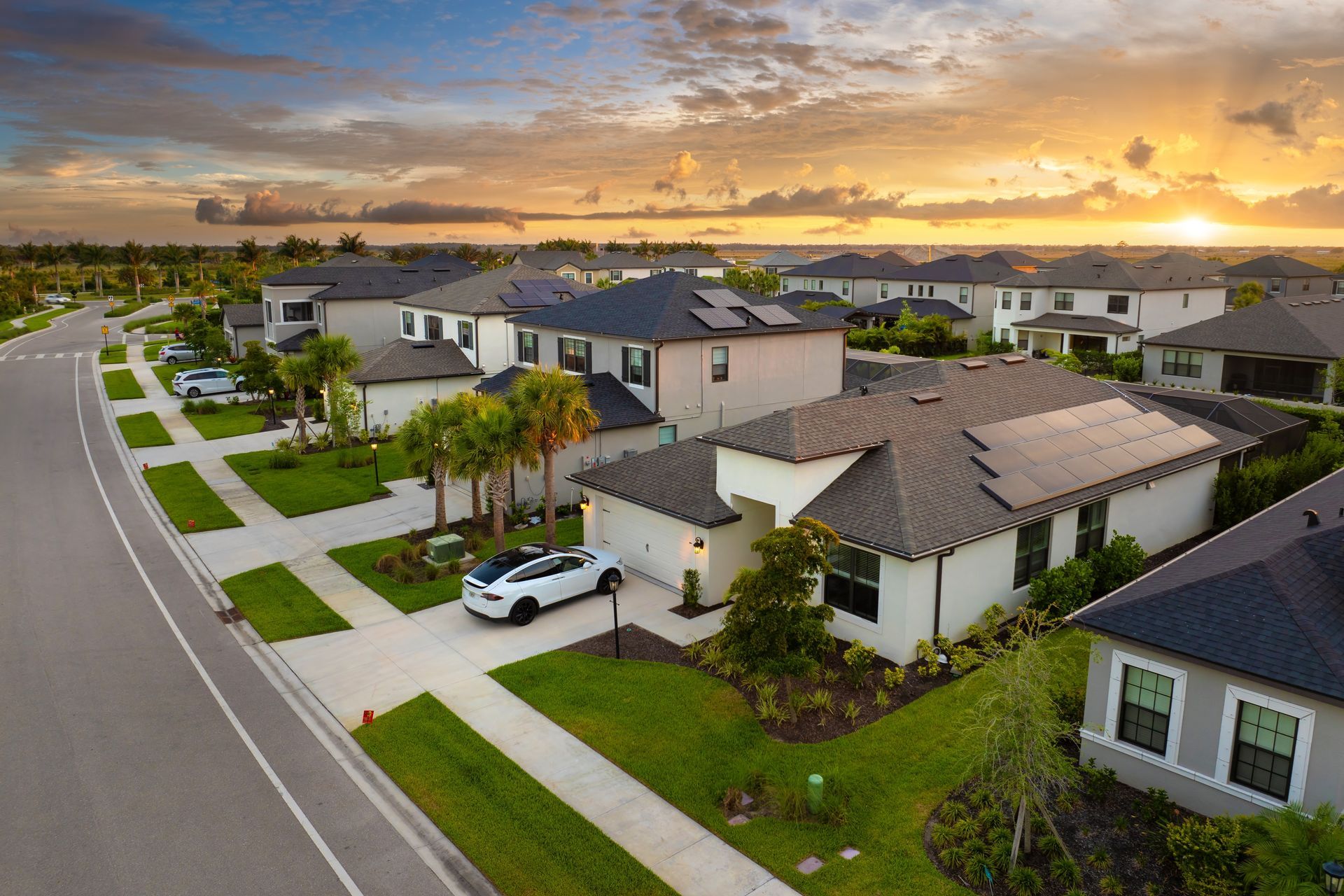 Aerial view of a suburban neighborhood at sunset with modern homes, sidewalks, and parked cars.