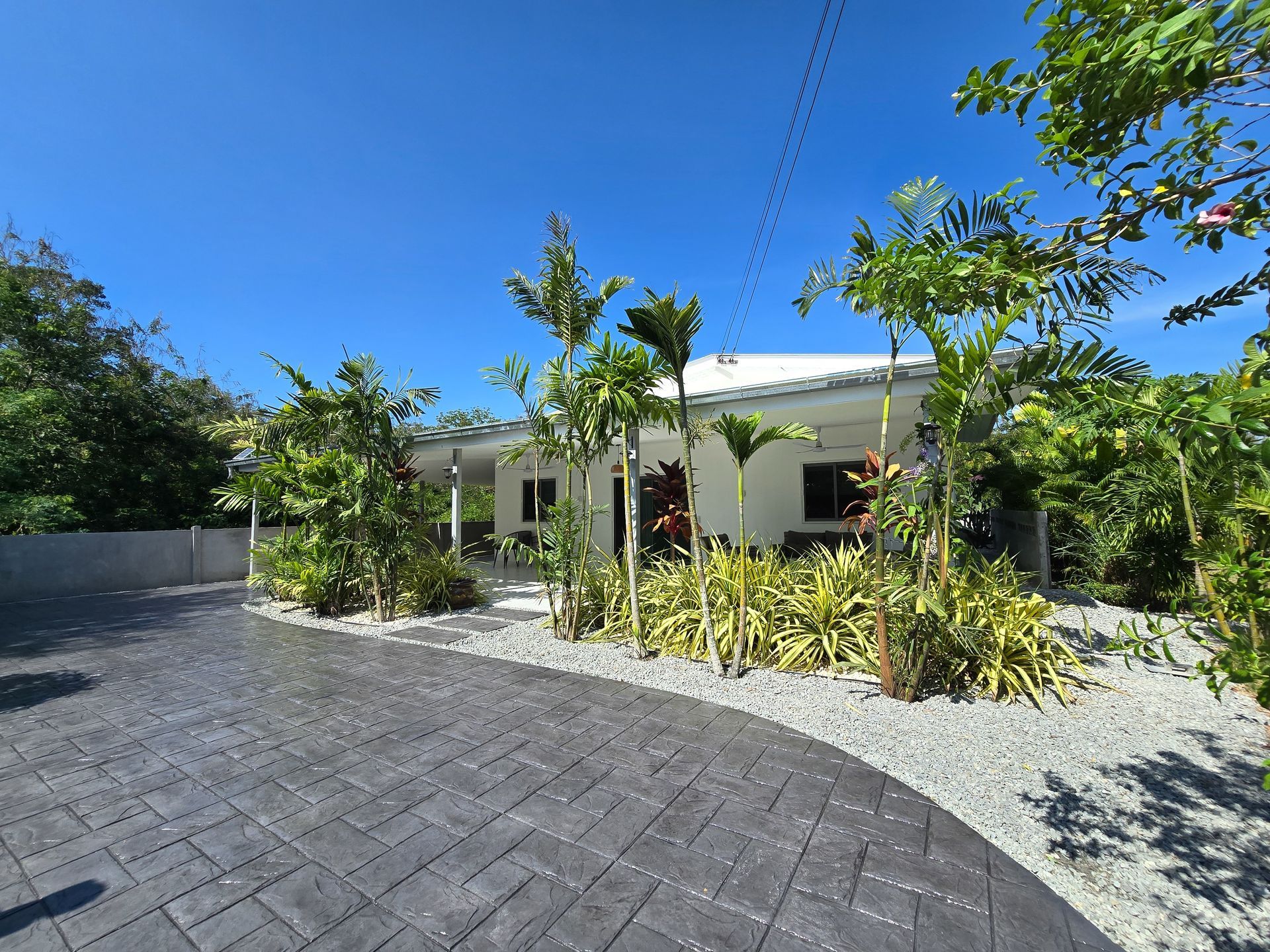 Single-story white house with a tiled driveway, tropical plants, and a bright blue sky
