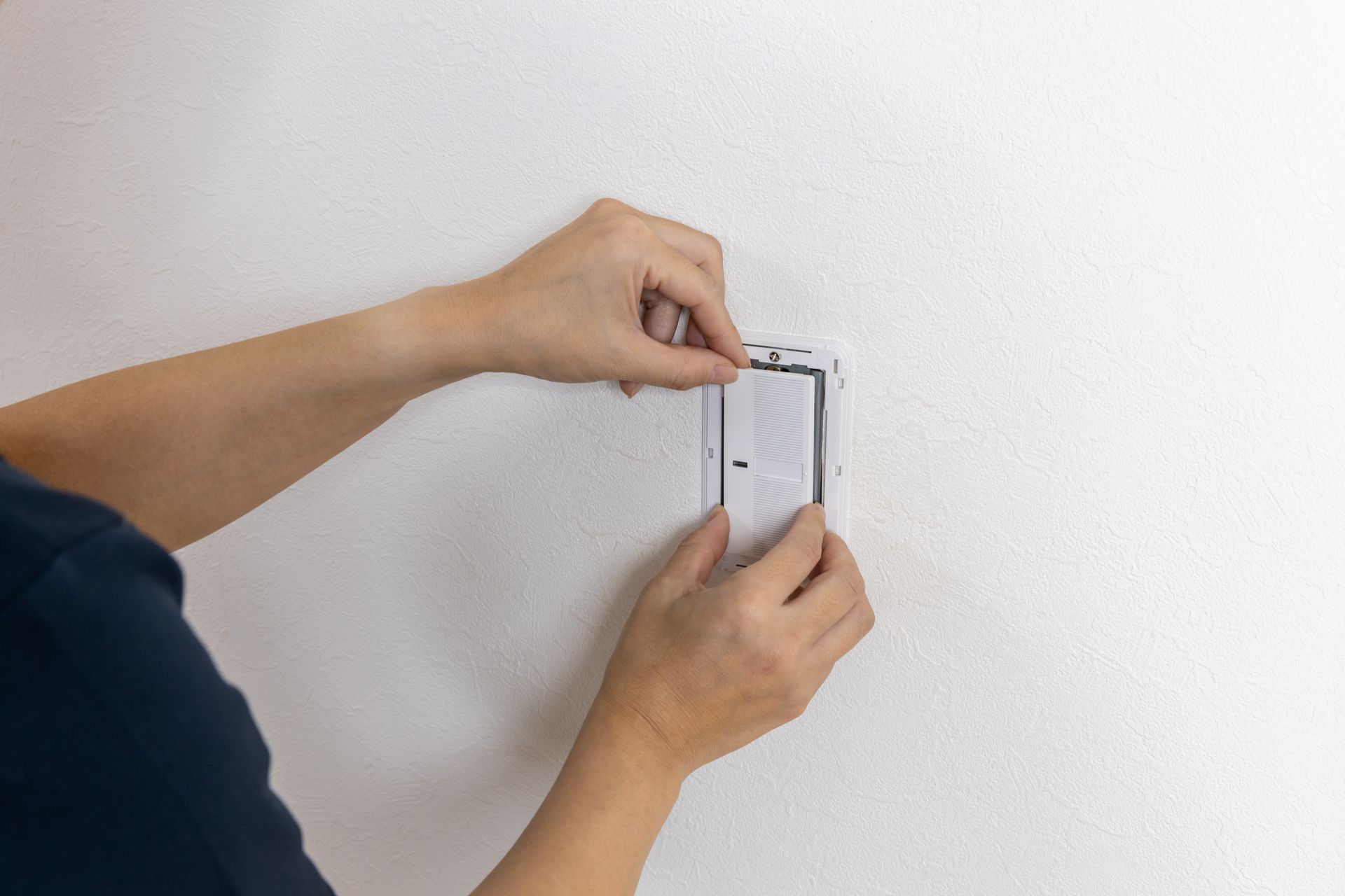 Hands installing a white wall-mounted device with a screwdriver.