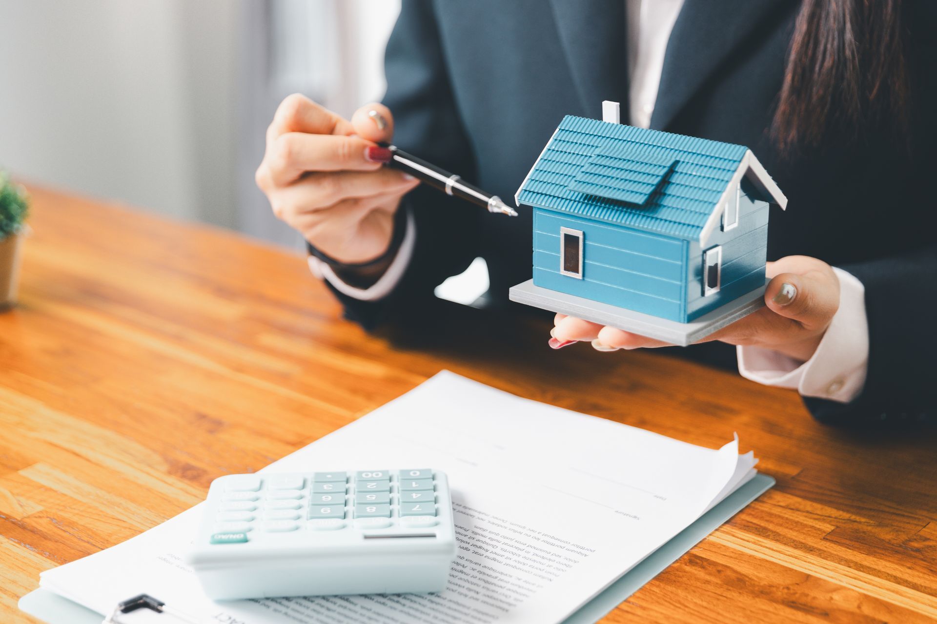 Person pointing at a blue model house beside calculator and paperwork on a desk